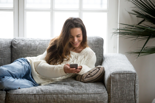 Happy,Young,Woman,Lying,On,Sofa,At,Home,Using,Smartphone,